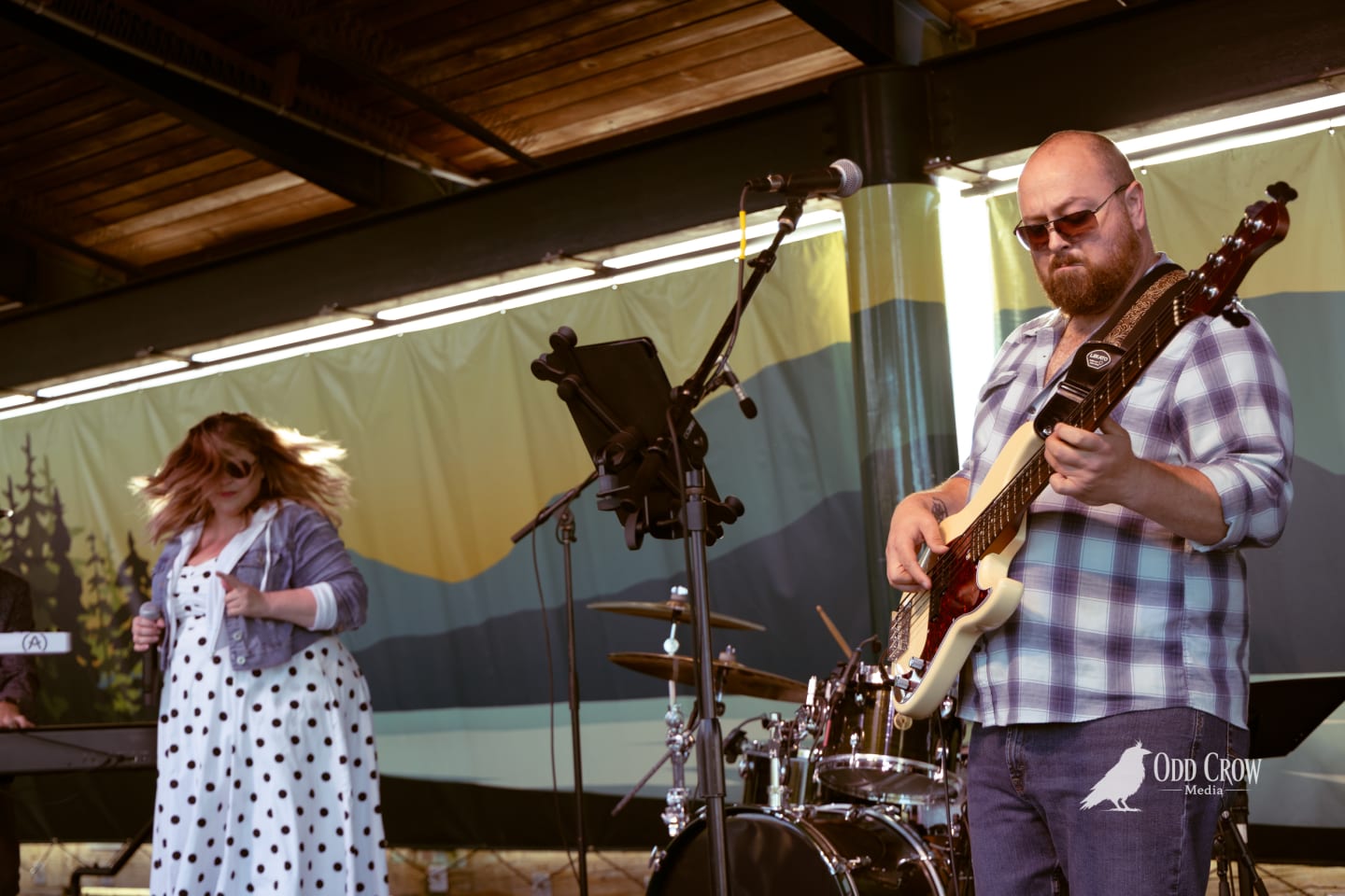 Guitarist in plaid shirt and singer in polka dot dress perform on an outdoor covered stage.