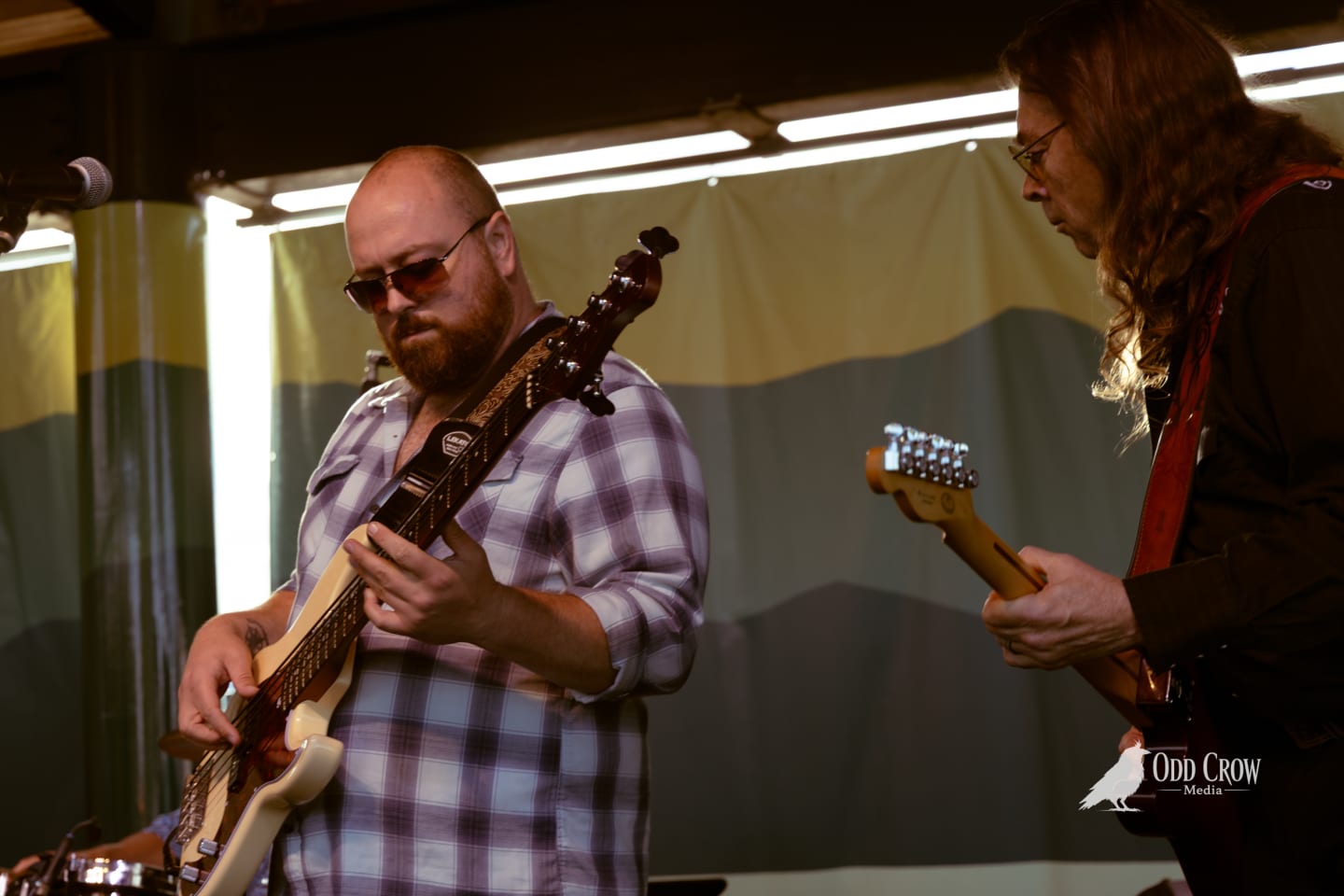 Two guitarists performing on stage, one bald with sunglasses playing electric guitar, at an outdoor pier venue.