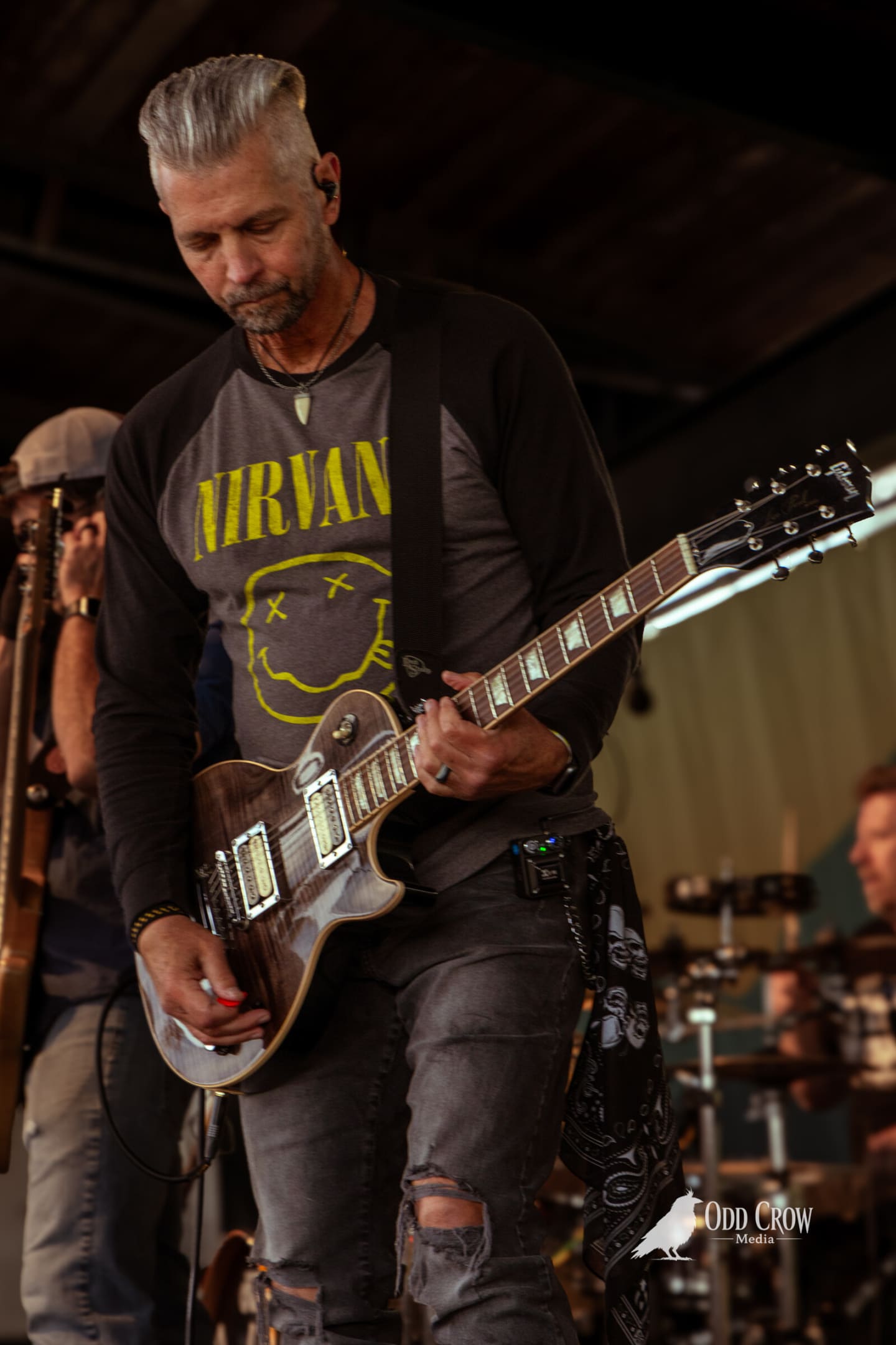Guitarist with silver hair in a Nirvana smiley shirt looking down at a worn Les Paul
