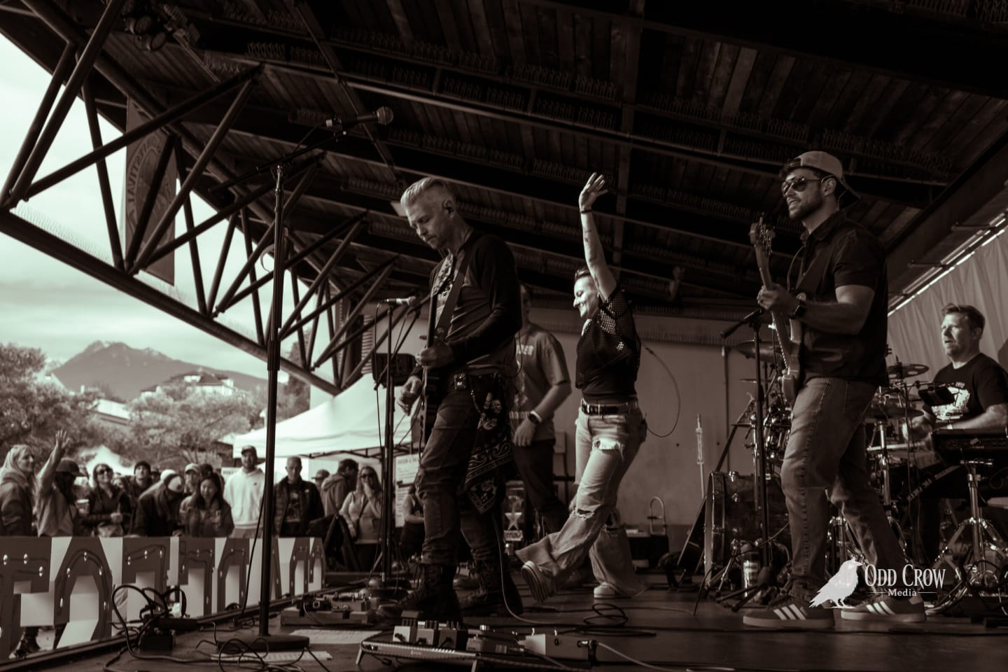 Black and white wide shot of the band mid-song under the stage roof, a vocalist's arm raised and a crowd watching from the left