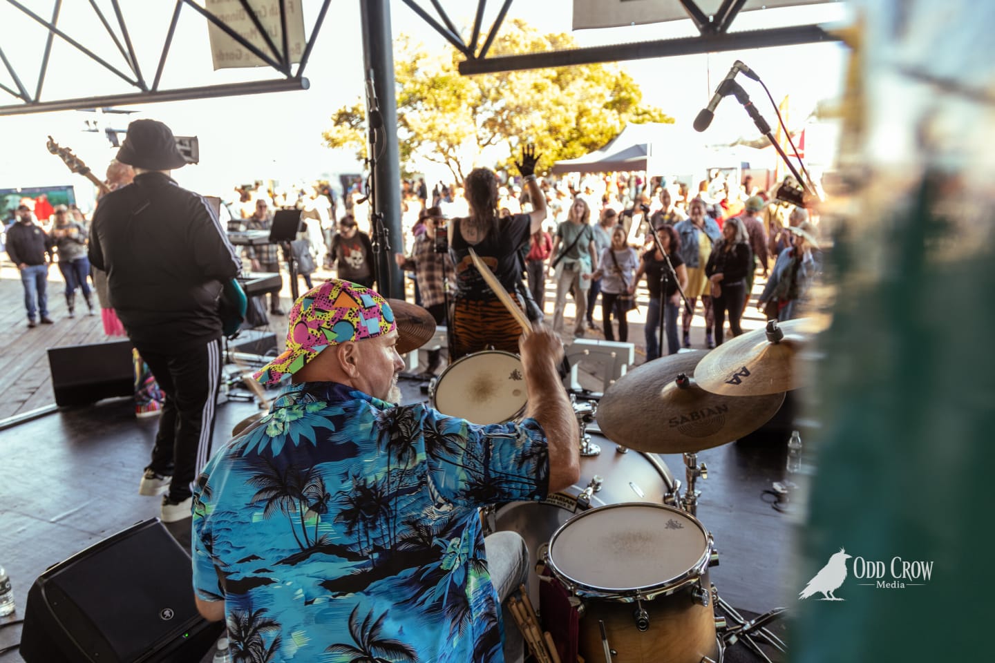 View from behind the drum kit, a drummer in a tropical shirt and bucket hat, looking out over a packed outdoor festival crowd