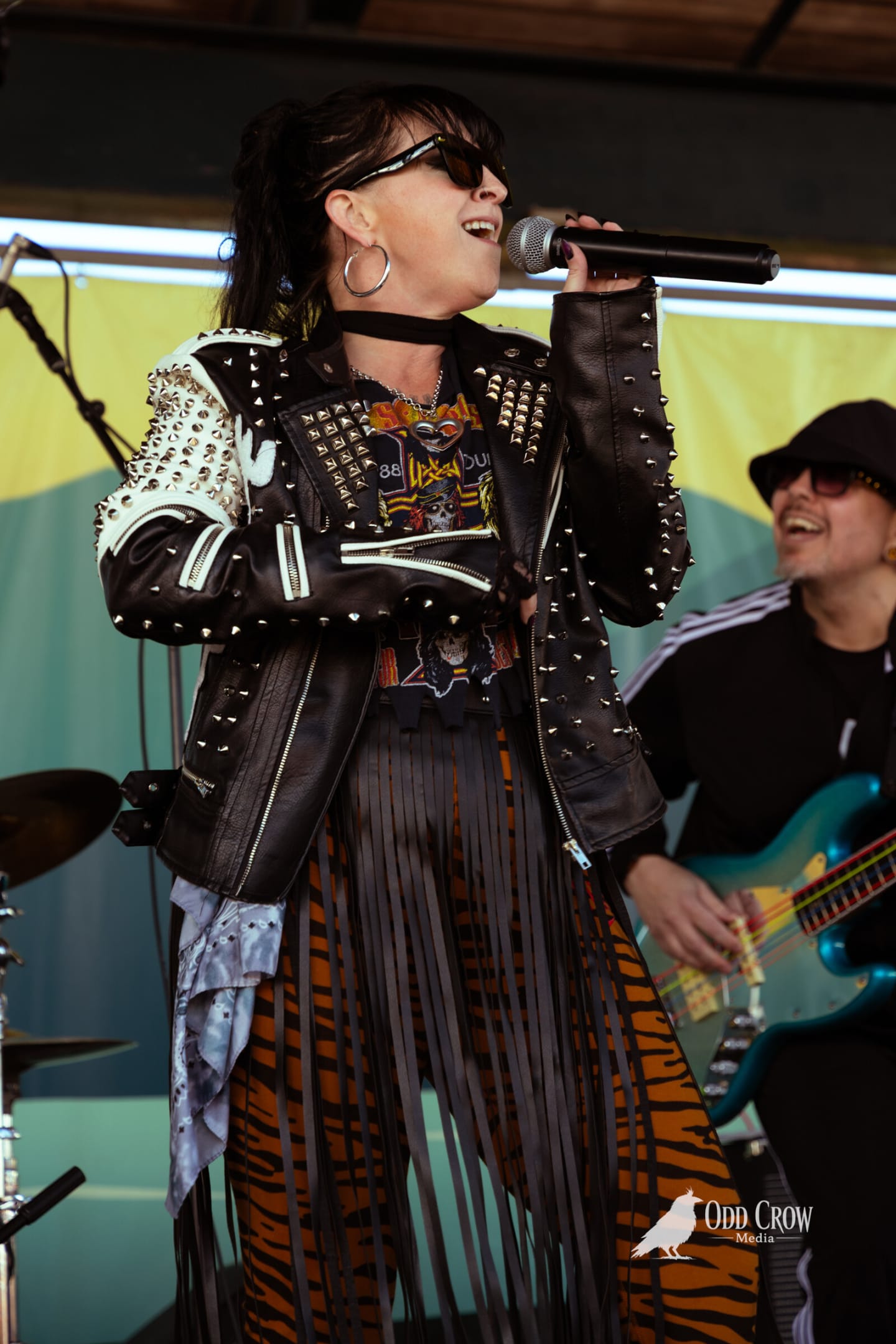 Female vocalist in a studded leather jacket and tiger-print skirt singing, bass player behind her