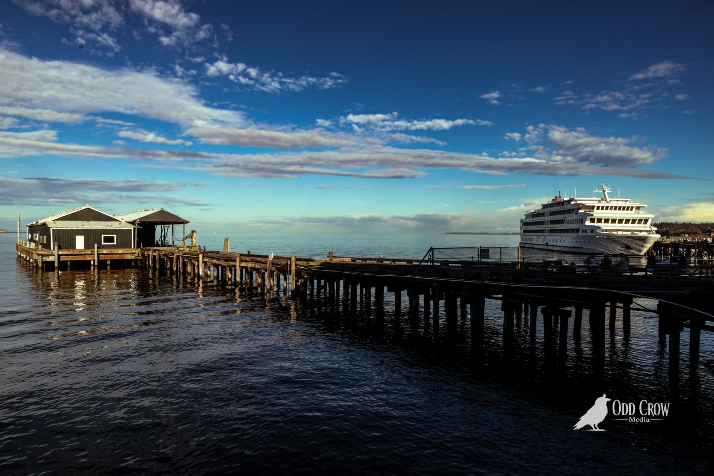 Wooden pier with small buildings and a cruise ship docked beyond it under a blue sky