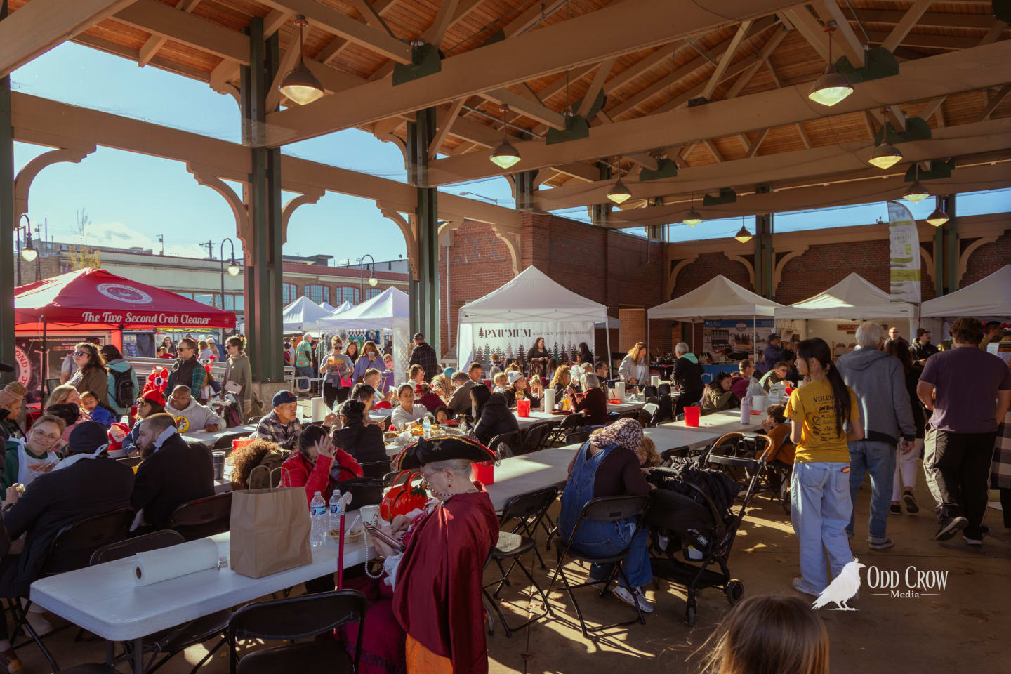 Families seated at long tables inside the covered festival pavilion, vendor booths and bright fall sunlight beyond