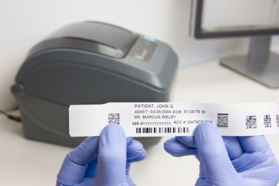 Gloved hands holding up Typenex Medical's hospital AdminBand with patient data; thermal printer in background.