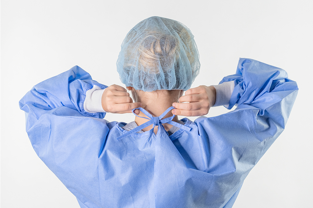 Typenex Medical PPE. Person wearing blue open-back surgical isolation gown, fastening tie closures around neck.