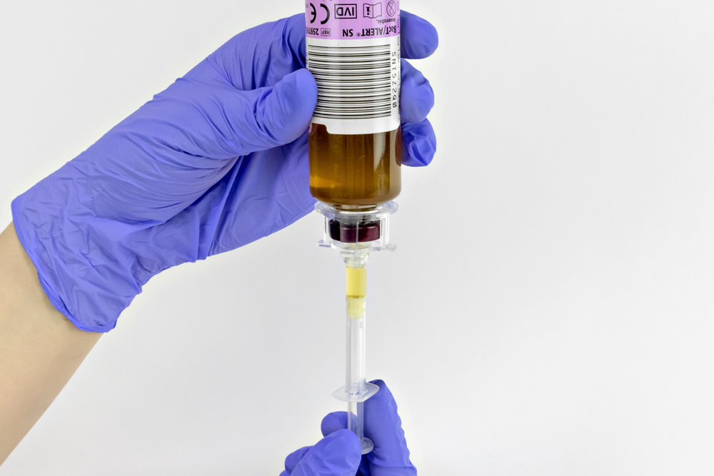 Gloved hand holding a culture bottle with CultureSafe culture bottle dispensing device affixed, using a syringe to draw a specimen sample.