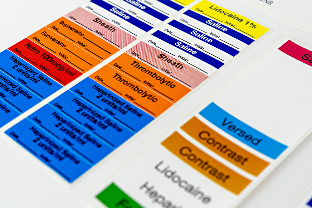 Stack of custom sterile labels in blue, orange, pink, and yellow displaying medications such as Saline, Heparin, Thrombolytics, Contrast, and Lidocaine with printed information fields.