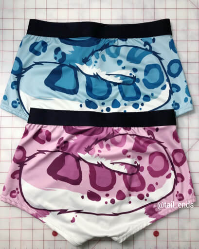 Two pairs of boxer shorts with black elastic waistbands, both depicting a snow leopard's tail and spots. One is blue and the other is magenta.