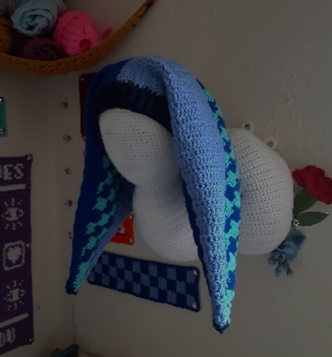 A crochet jester hat in various shades of blue that sits on a white wig head with a very long and curled neck. the wig head also has a crocheted cover.