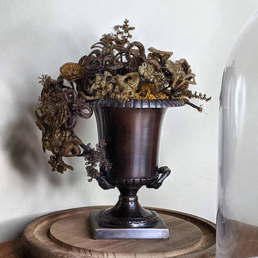 A vase filled with flowers in various shades of brown that are spilling over the left side. Each flower is made of human hair.