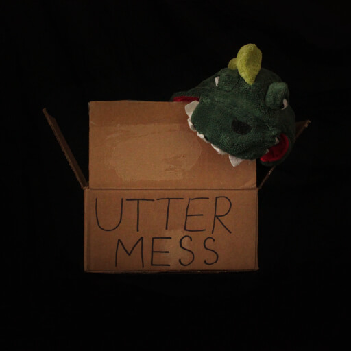 A cardboard box with the head of a green dinosaur onesie spilling out sits in a black void. On the box UTTER MESS is written in black marker.