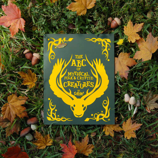 Picture of a coloring book cover, dark green color with yellow text that reads: The ABC of Mythical, Folk & Cryptid Creatures. The book is laying over a grass area surrounded by fall leaf and mushrooms.