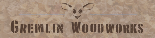 A banner image with the words "Gremlin Woodworks" in dark brown in a block stencil font. Centered above the words is a simple line drawing of a mischievously smiling face with large pointed ears. The background of the image is a washed out macro photo of a pile of multi-colored wood shavings.