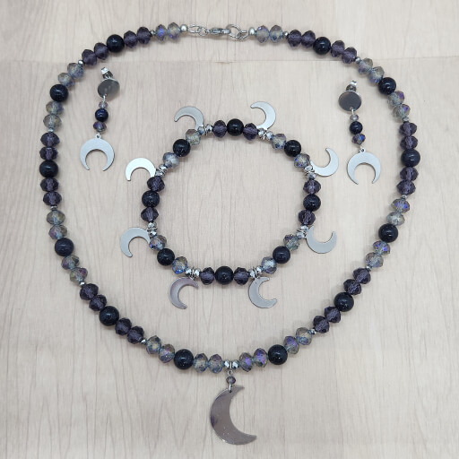 A set of necklace, bracelet and earrings, made with beads in shades of dark purple and blue, with silver toned half-moon charms.