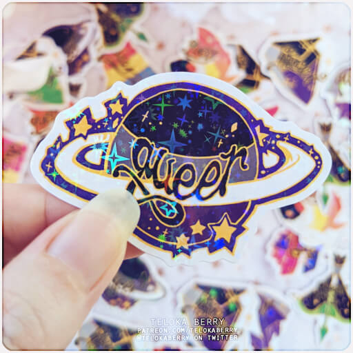 Photograph of a die-cut sticker with a starry holographic effect depicting a ringed planet in the colours of the asexual flag with the word "queer" written over the top in script font