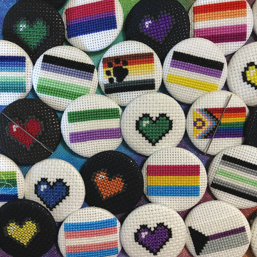 Hand-stitched cross stitch 1.5” buttons in a variety of Pride flag designs, including non-binary, demisexual, agender, and transgender, and solid color hearts.