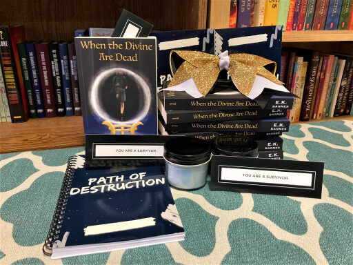 A stack of six books labeled "When the Divine Are Dead" with the author's name "E.K. Barnes" on the side, as well as the number 1. A large yellow, white, and black bow sits upon the stack. Behind it are two navy-colored journals. Next to the stack is the same book, but with the cover showing. The cover has a boy running inside a crystal ball. Above it is the title, "When the Divine Are Dead." Below the book is a black and white bookmark that says, "You are a survivor." In front of the bookmark is another navy-colored journal that reads, "Path of Destruction." Next to the journal are two light blue candles in small jars. Another survivor bookmark is in front of one of the candles. Behind everything is a bookshelf filled with various books.