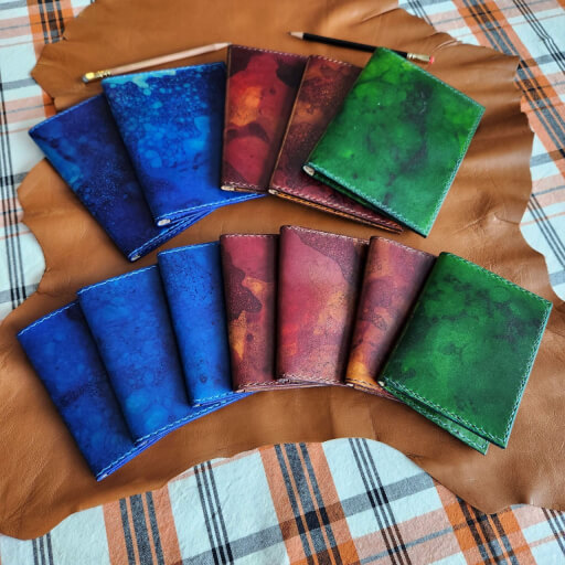 The image is of several notebook covers fanned out in two rows. Abovethe top row are two pencils. The top row is five larger notebook covers (two blue, two brown, one green), and the lower row is seven smaller notebook covers (three blue, three brown, one green). The color on all the notebook covers have a bubble and mottled finish. The covers and pencils lay on small brown leather hide, which is on an orange and black plaid tablecloth