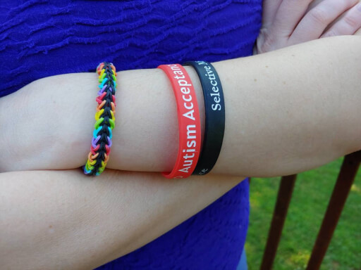 A close up on the wrist of a person whose arms are crossed. The person is wearing a black and rainbow colored rubber band bracelet, a red autism acceptance silicone wristband, and a black selective mutism silicone wristband.