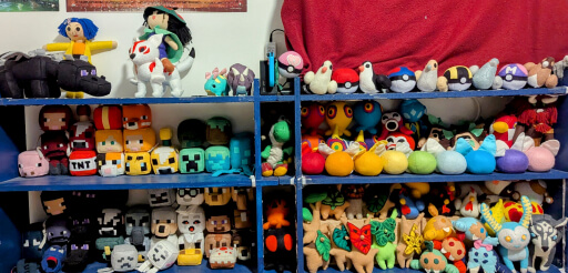 A long low book shelf painted dark blue, filled with colorful plush characters and animals. On the shelves are: top left, a Minecraft ender dragon, a Coraline doll, and an okami Ameterasu and Issun. Top right, a variety of pigeons, with Pokeballs between them. Middle and bottom left, Minecraft blocks of all kinds, from characters to animals to enemies to crafting tables. Middle and bottom right, Characters, creatures, and items from the legend of Zelda series. Some of them, like the fairies, are arranged in rainbow order.