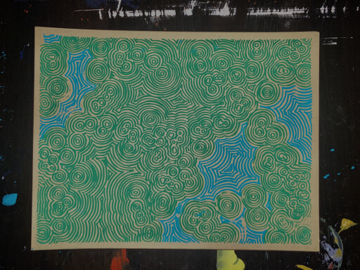 A small 8.5x11 two color screenprint on brown cardstock. There are emanating lines in green and blue giving the appearance of a semi-abstracted topographical map. The print is lying on an inkstained black table.