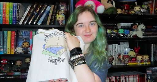 White girl with green hair sitting in front of a black bookcase filled with books and funky pops. She is holding a white tote bag with the image of a stack of three books on it. The bottom book is a pastel blue, the middle is pastel green and the top is an open book. Around the image are the words 'just one more chapter' in a black and white font. She is hugging the bag to her left, next to her head, the bag itself is full of books, which are not visible.