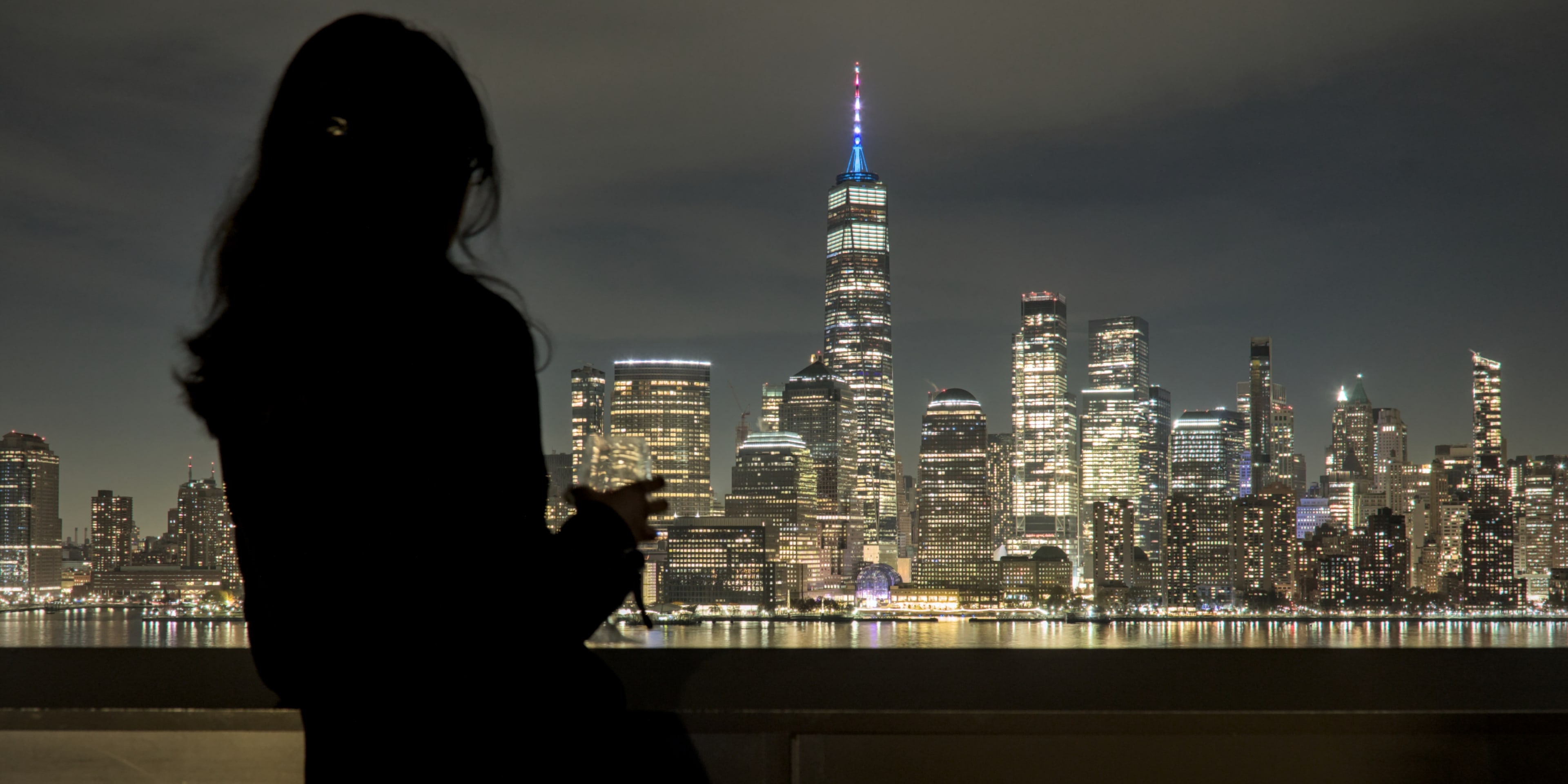 woman in a winter coat looking out at the nighttime NYC skyline