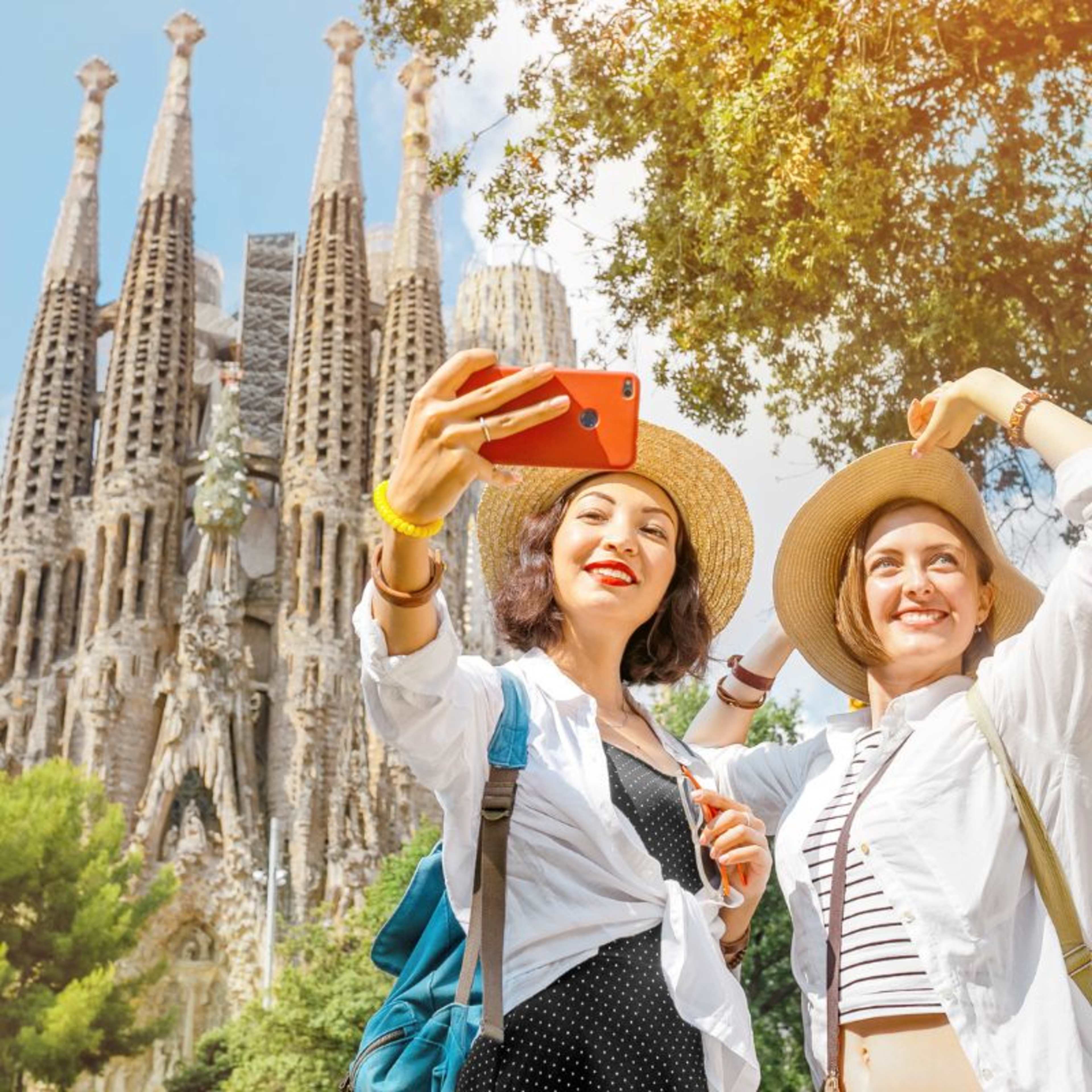 Two ladies in straw hats take a selfie in the sunshine in front of the Sagrada Familia, Barcelona