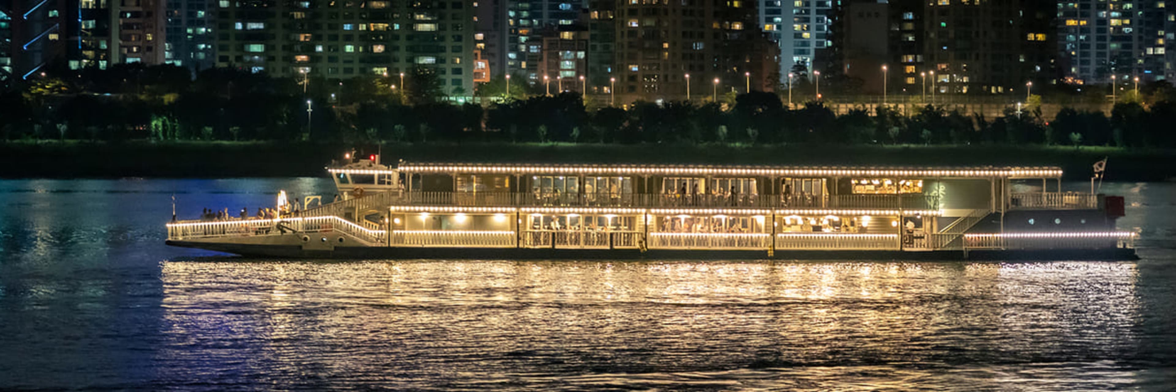 The E-land Han River cruise boat illuminated against Seoul's city skyline at sunset.