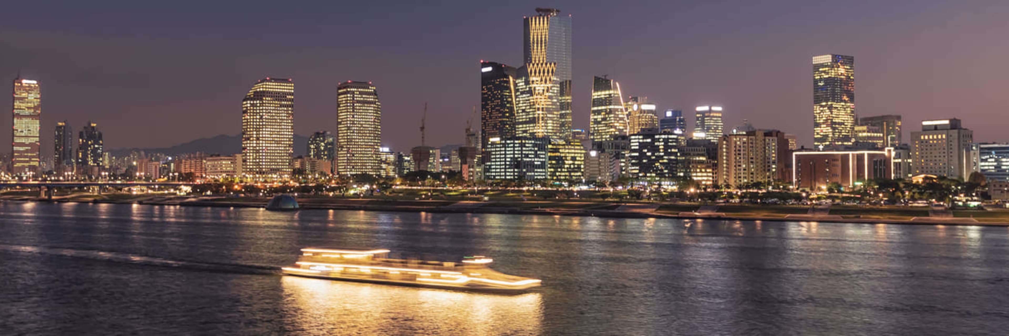 The E-land Han River cruise boat illuminated against Seoul's city skyline at sunset.