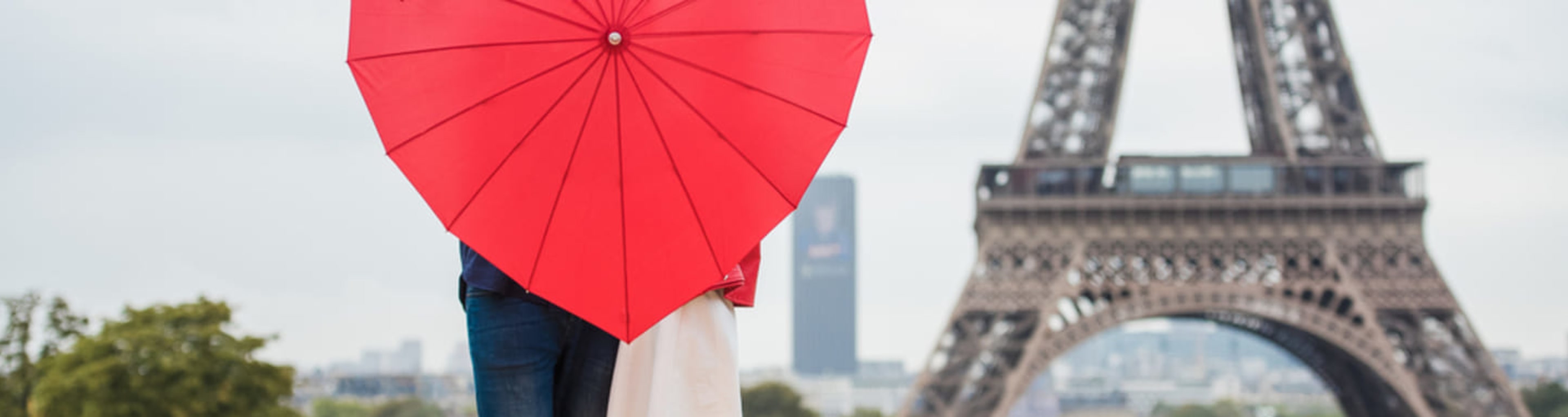 Couple holding a heart-shaped umbrella in front of the Eiffel Tower.