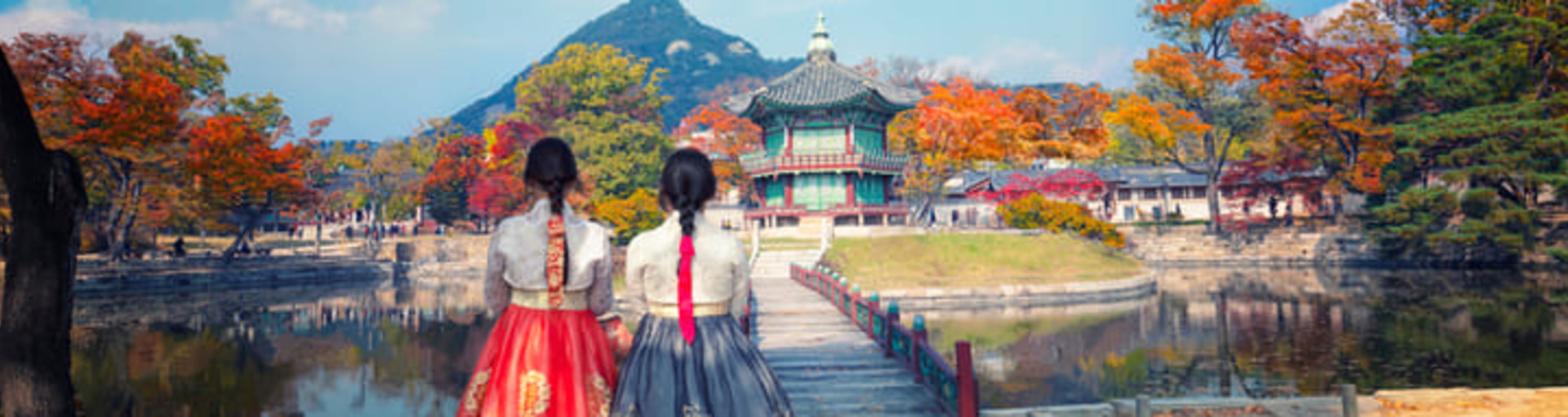 Friends in traditional Korean dress gazing at Gyeongbokgung Palace beneath red autumn leaves.