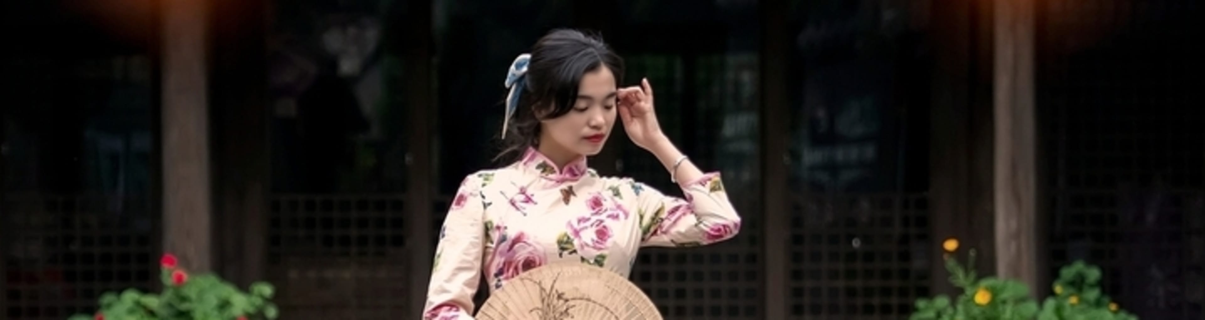 Woman in traditional floral cheongsam dress, holding a paper fan.