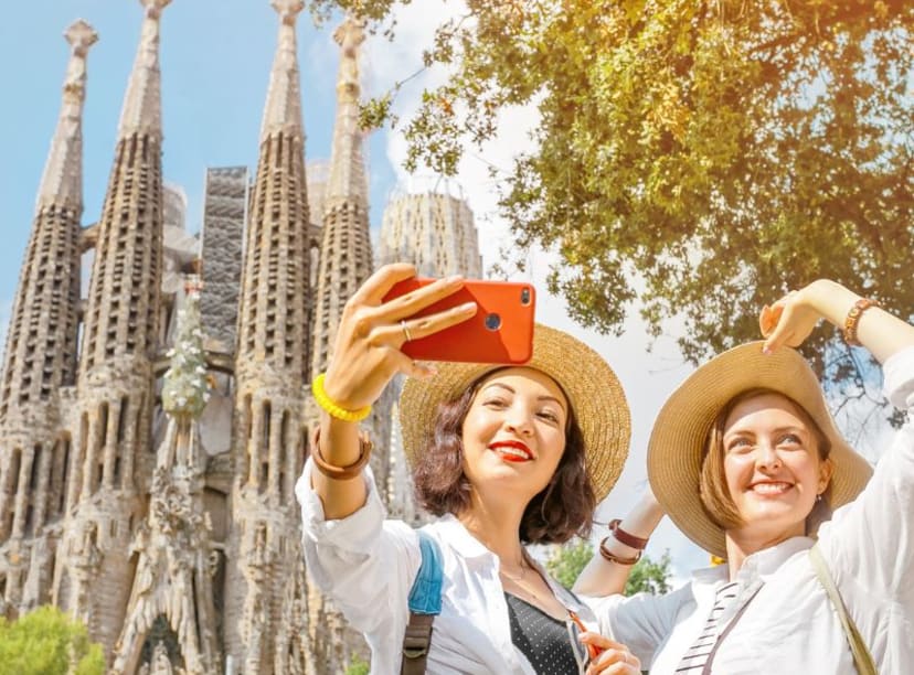 Two ladies in straw hats take a selfie in the sunshine in front of the Sagrada Familia, Barcelona