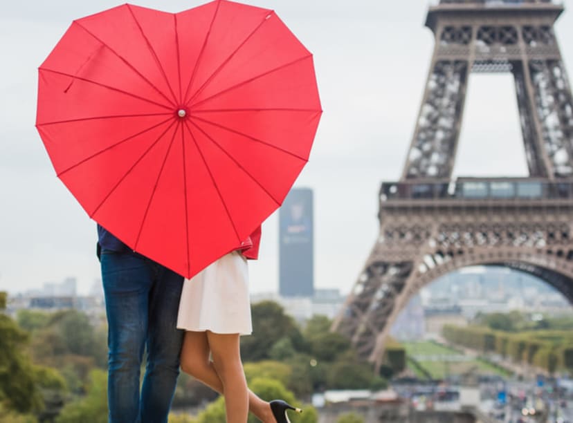 Couple holding a heart-shaped umbrella in front of the Eiffel Tower.