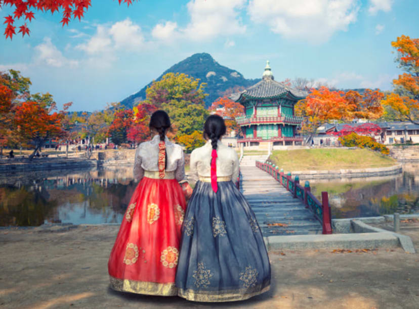 Friends in traditional Korean dress gazing at Gyeongbokgung Palace beneath red autumn leaves.