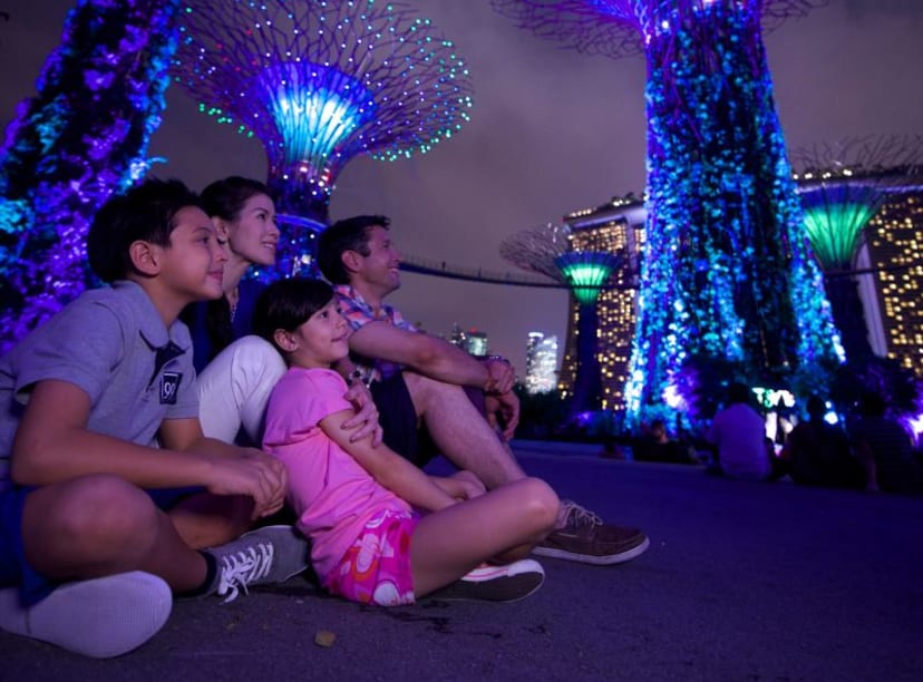 Family of four sit on ground after dark looking up at three Singapore supertrees lit in blue