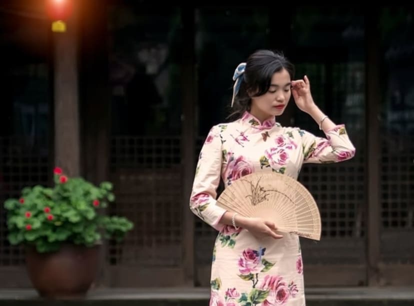 Woman in traditional floral cheongsam dress, holding a paper fan.