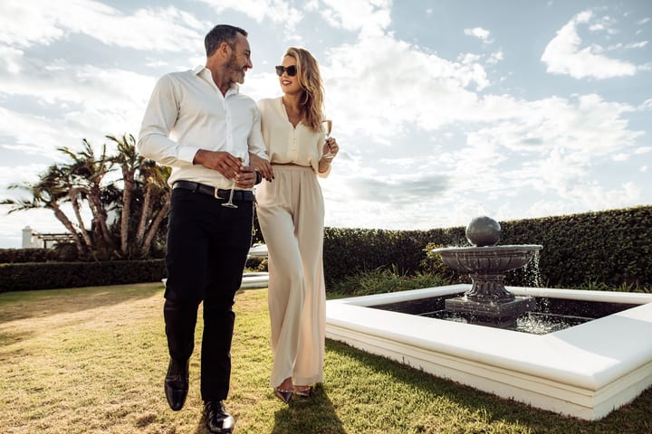 Pareja adinerada en un jardín con árboles y fuente.