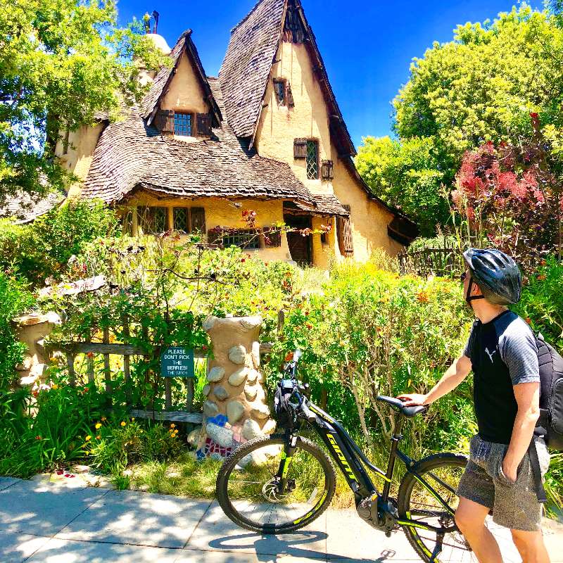 Image of Building, Cottage, House, Housing, Wheel, Helmet, Female, Girl, Person, Teen, Bicycle, Vehicle, Shorts, Outdoors, 