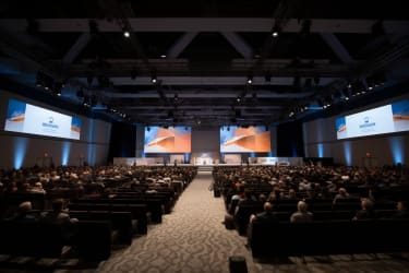 A professional conference in a spacious hall with high ceilings and large windows letting in ample natural light. The hall is filled with rows of chairs, facing a stage with a podium and a screen. The stage is adorned with elegant decorations and banners with the logos of various companies and organizations sponsoring the event. On the stage, a speaker is delivering an engaging talk, holding a microphone and using a laser pointer to highlight key points on the projected slides. The audience members, dressed in casual attire, are taking notes on their tablets and laptops, engaged and attentive. The atmosphere is serious yet vibrant, with a sense of shared enthusiasm and intellectual curiosity permeating the space. The composition emphasizes the grandeur of the hall, with the rows of chairs converging towards the stage, creating a sense of focus and direction. The color palette is subdued, with shades of blue, gray, and black predominating, lending an air of professionalism and sophistication. The image is shot with a Canon EOS R6 camera, using a Canon EF 24-70mm f/2.8L II USM lens at 50mm, ISO 400, f/4.0, 1/125s, --v 5 --ar 3:2