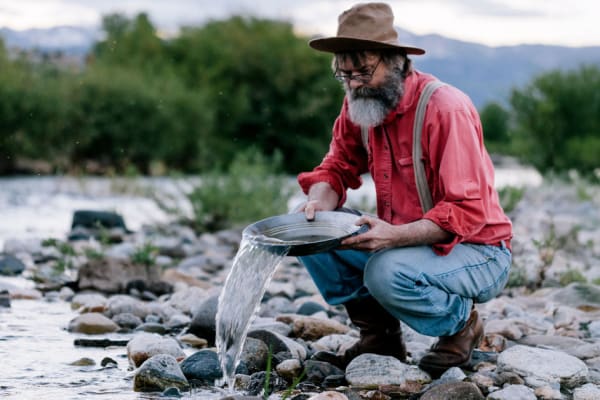 A prospector, crouched at the edge of a river, panning for gold