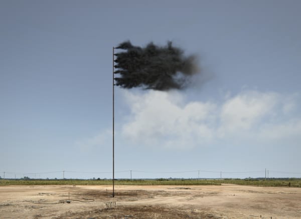 A pole with dark smoke coming out of the top, creating a flag-like shape, stands in a flat, sandy area.
