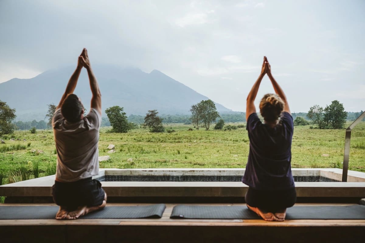 Un huésped disfruta de una actividad de bienestar en Kwitonda Lodge, rodeado de exuberante vegetación y tranquilos paisajes naturales, con los picos volcánicos cubiertos de niebla del Parque Nacional de los Volcanes de Ruanda visibles en el fondo. El entorno sereno combina lujo, relajación y vistas impresionantes del paisaje montañoso icónico del parque.