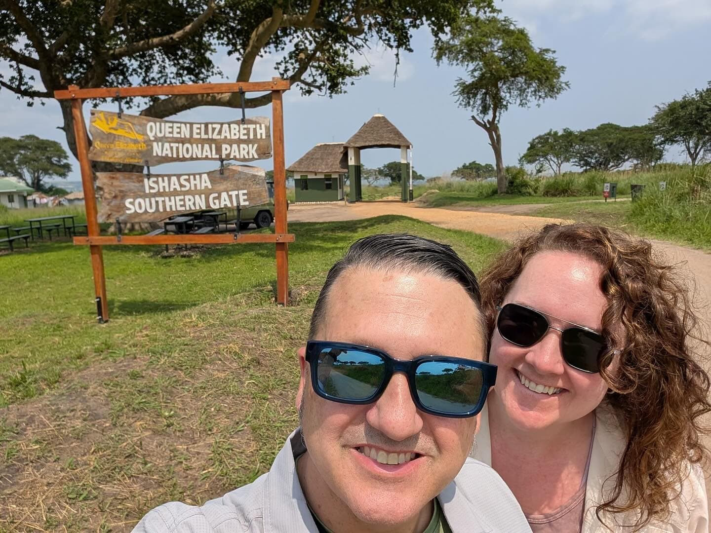 A couple smiling beside the Ishasha sector entrance sign of Queen Elizabeth National Park