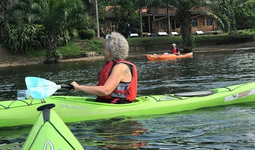 A woman in a life jacket kayaking on a lake with a lodge on the shore