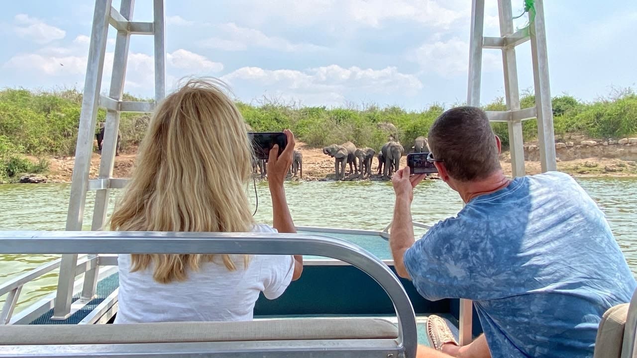 Two guests on a boat photographing animals on the shore of the Kazinga Channel