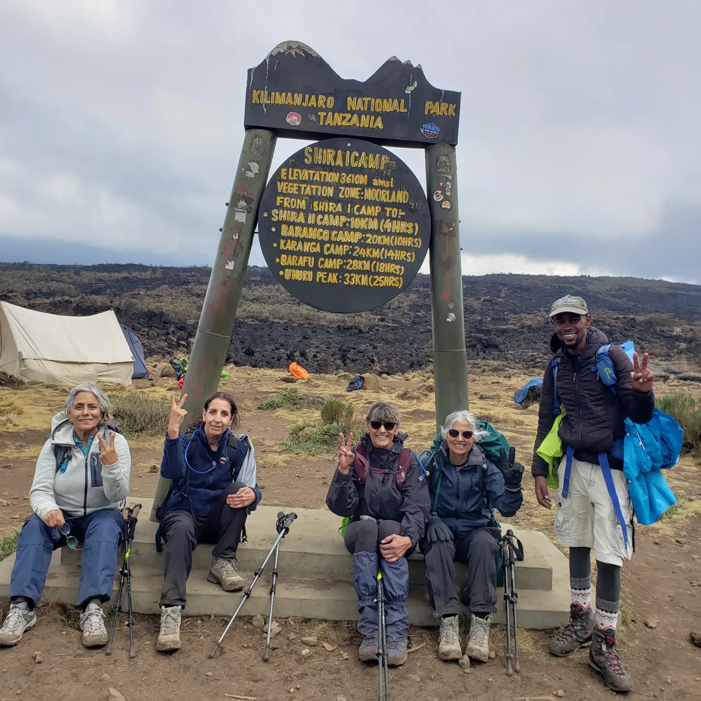 Five hikers resting beside a park signpost at a camp on Mount Kilimanjaro
