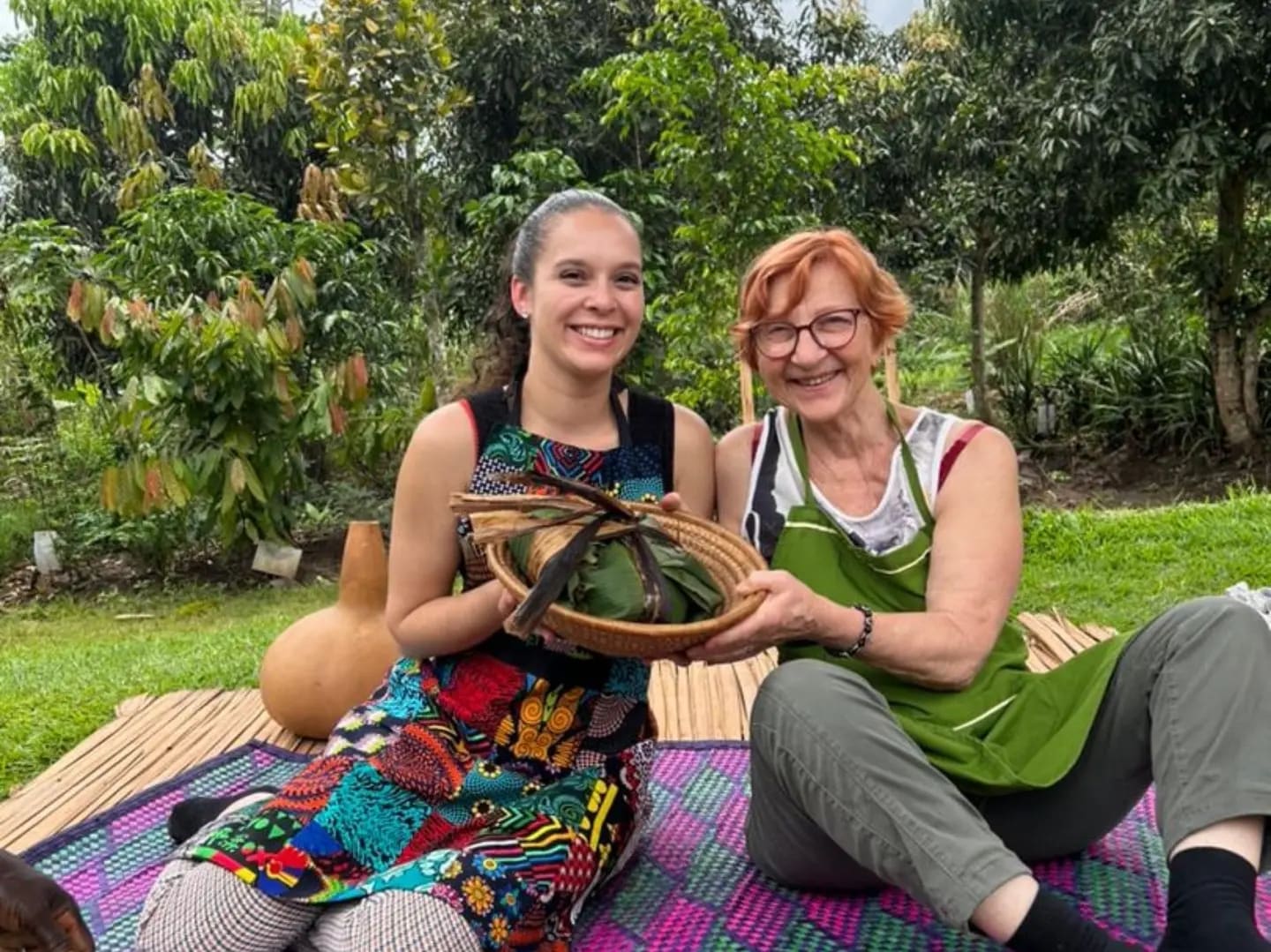 Two women holding a basket of local food on a picnic mat in a garden